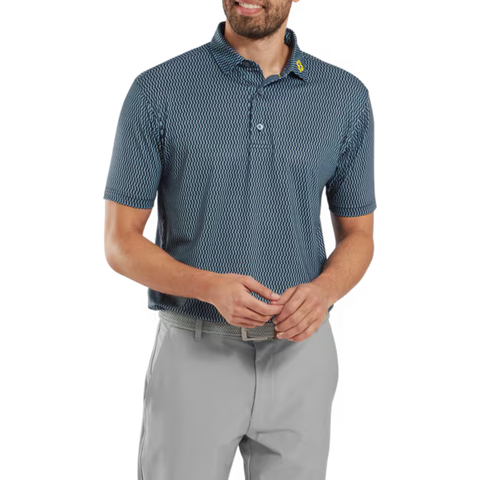A man, smiling with hands loosely clasped, wears a FootJoy Men’s Wiggle Print Lisle Polo in Navy/White and light gray pants while standing against a white background.