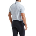 A man in a light gray patterned FootJoy Scottish Town Mens Print Polo and dark trousers stands facing away with hands in his pockets. The moisture-wicking polo has a small black logo near the collar.