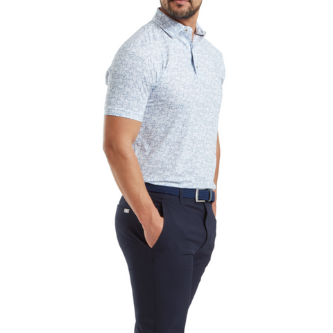 A man in a FootJoy Scottish Town Mens Print Polo, featuring a light blue pattern, with navy pants and a blue belt stands with hands in pockets against a white background.