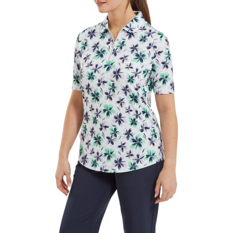 A woman wears a FootJoy Women's 1/4 Zip Short Sleeve Floral Shirt—a white, moisture-wicking golf top with blue and green floral prints—paired with dark pants. She stands facing slightly left, arms relaxed at her sides.