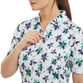 A woman wears a FootJoy Women's 1/4 Zip Short Sleeve Floral Shirt in Lavender/Mint/Teal, partially unzipping the front; only the lower part of her face and upper body are visible.