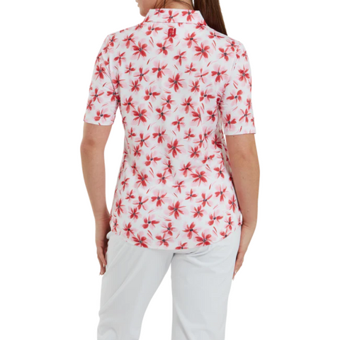 A person faces away, wearing a FootJoy Women's 1/4 Zip Short Sleeve Floral Shirt with a red and pink floral pattern and white pants. The collared, moisture-wicking shirt from FootJoy pairs perfectly with their hair pulled to one side.