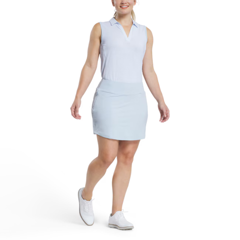A woman in a sleeveless light blue collared shirt, FootJoy Women’s Aerial Performance Knit Skort in Skyway Blue, and white shoes walks forward against a plain white background.
