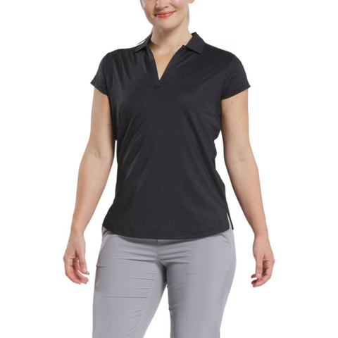 A woman wears a FootJoy Women's Cap Sleeve Print Shirt in Black Animal Print, standing against a white background and smiling slightly, with her arms relaxed at her sides and paired with light gray pants.
