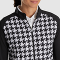 A woman wears a FootJoy Womens Full Zip Houndstooth Mid Layer Jacket in black and white, featuring a bold houndstooth pattern on the front and black sleeves over a white collared shirt. Only her lower face and upper body are visible.