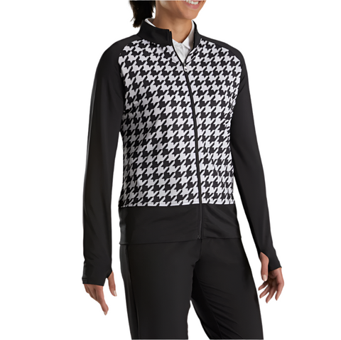 A person models the FootJoy Womens Full Zip Houndstooth Mid Layer Jacket in black and white, featuring a houndstooth front and solid black sleeves and sides, paired with black pants and smiling while facing slightly to the side.