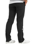 A person wearing black FootJoy Women's HydroLite V2 Rain Trousers with a white logo near the left ankle, paired with white sneakers and a white top, stands facing away from the camera with their right hand in their pocket.