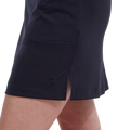 Close-up of a person wearing the FootJoy Womens Interlock Stretch Skort in navy, featuring a small side slit and subtle stitched FootJoy logo near the hem, shown from mid-thigh down.