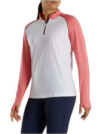 A woman wears the FootJoy Women's Sun Protection Shirt in White/Coral, featuring long light coral sleeves, a white body, and front half-zip, paired with dark pants. This UPF 50+ FootJoy golf shirt is perfect for outdoor style and sun protection.