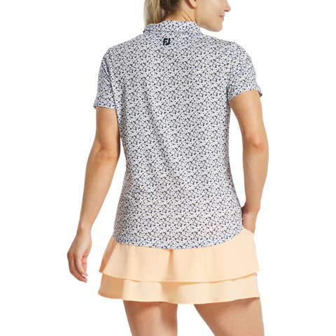 A woman is seen from behind wearing a FootJoy Women's Floral Print Short Sleeve Shirt with a white collar, paired with a light peach tiered skirt, standing against a plain white background.