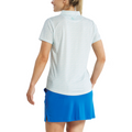 A woman faces away, wearing a FootJoy Women's Floral Print Short Sleeve Shirt—a light-colored collared top—paired with a bright blue skirt with side slits.