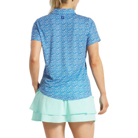 A woman with blonde hair pulled back is seen from behind wearing a FootJoy Womens Floral Print Short Sleeve Shirt in blue and a light turquoise layered skort, standing against a plain white background.