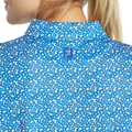 Close-up of a woman in the FootJoy Womens Floral Print Short Sleeve Shirt, featuring a white speckled pattern and blue embroidered FJ logo near the collar. Her blonde hair is tied back.