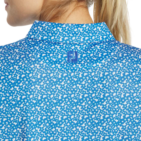 Close-up of a woman in the FootJoy Womens Floral Print Short Sleeve Shirt, featuring a white speckled pattern and blue embroidered FJ logo near the collar. Her blonde hair is tied back.