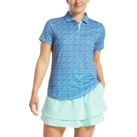 A person wearing a FootJoy Womens Floral Print Short Sleeve Shirt and a light blue tiered skirt stands with one hand in their pocket, smiling.