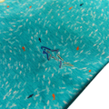Close-up of Greg Norman Men’s ML75 Shark Frenzy Print Polo in Aquamarine, showing blue fabric with small white, black, and orange shark prints plus an embroidered blue shark—ideal detail for a stylish, moisture-wicking golf polo.