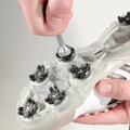 Someone uses the Masters Deluxe Spike Wrench by Masters to tighten or install a cleat spike on the sole of a white athletic shoe featuring several black cleat fittings.