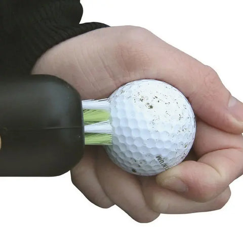 A hand uses the Masters Water Bottle Club Cleaner by Masters, featuring white bristles, to scrub a dirty golf ball clean against a plain white background.