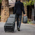 A person in black pants and a jacket walks on a paved path, pulling a large wheeled golf travel bag and carrying the Ecco Ping Shoe Bag, with stone buildings in the background.