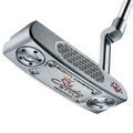 A close-up of the Scotty Cameron 2025 Studio Style Newport Plus Putter shows engraved branding, a precision milled silver head, a textured face insert, and red, black, and gold accents.