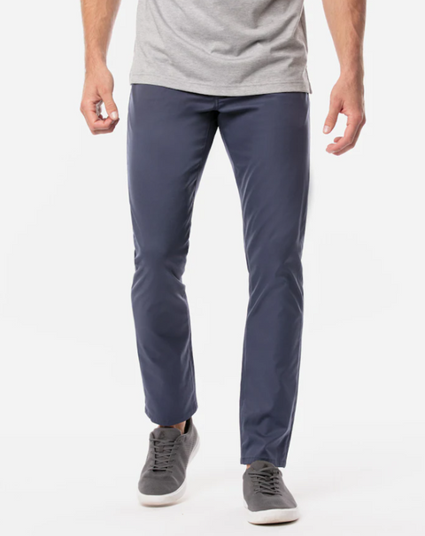 A person stands against a plain white background wearing Travis Mathew Men's Open to Close Pants in Heather/Navy, paired with a light grey t-shirt and grey sneakers.