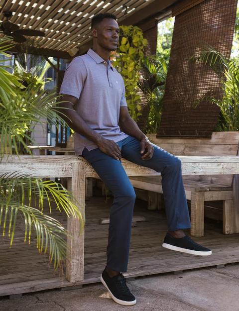 A man in a light purple polo, Travis Mathew Men's Open to Close Pants in Heather/Navy, and black sneakers sits on a rustic wooden table outdoors amid lush greenery and wood structures.