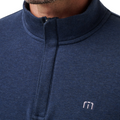 Close-up of a person wearing the Travis Mathew Cloud 1/4 Zip 2.0 Men’s Pullover in Heather Blue, showcasing its mock neck and small embroidered logo on the left chest. Only part of the lower face and neck is visible.