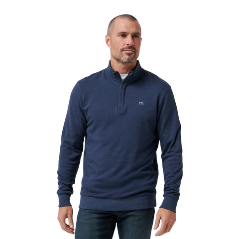 A man with a trimmed beard and short hair wears a Travis Mathew Cloud 1/4 Zip 2.0 Men’s Pullover in heather blue and dark jeans, standing against a plain white background.