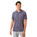 A man with short dark hair stands against a white background, smiling in a Travis Mathew Featherweight Flourish Men’s Polo—navy blue with red and white floral pattern—paired with light gray pants and a wristwatch on his left wrist.