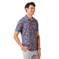 A man smiles in the TravisMathew Featherweight Flourish Men’s Polo—a blue, short-sleeve button-up with a red and white floral pattern—paired with light gray pants. He stands against a white background with one hand in his pocket.