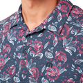 A close-up of a person wearing the Travis Mathew Featherweight Flourish Men’s Polo, a navy blue shirt with a red and white floral pattern, buttons, and an embroidered logo; the person’s face is not visible.