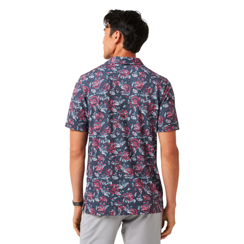 A man stands with his back to the camera, wearing a Travis Mathew Featherweight Flourish men’s polo featuring a pink and white floral pattern and light gray pants.