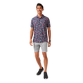 A man wearing the Travis Mathew Featherweight Flourish Men’s Polo in dark color, paired with light gray shorts and black sneakers, smiles while standing against a white background.