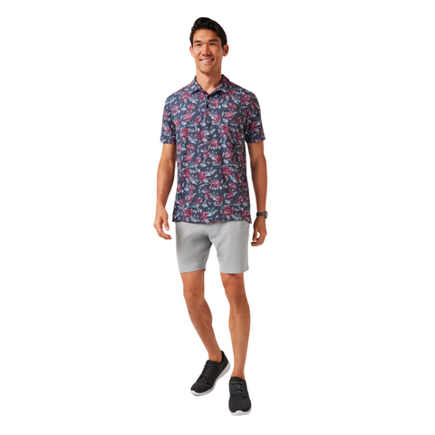 A man wearing the Travis Mathew Featherweight Flourish Men’s Polo in dark color, paired with light gray shorts and black sneakers, smiles while standing against a white background.