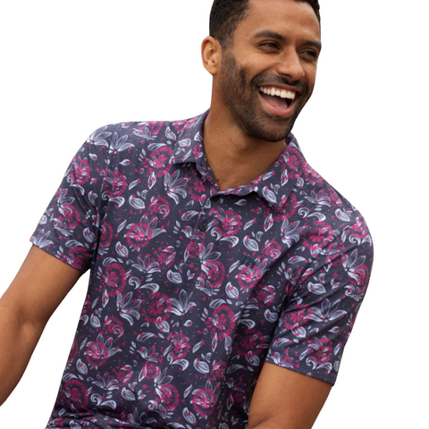 A smiling man wears the TravisMathew Featherweight Flourish Men’s Polo, a short-sleeved, navy-blue shirt with a pink and purple floral pattern, sitting relaxed and looking slightly to the side.