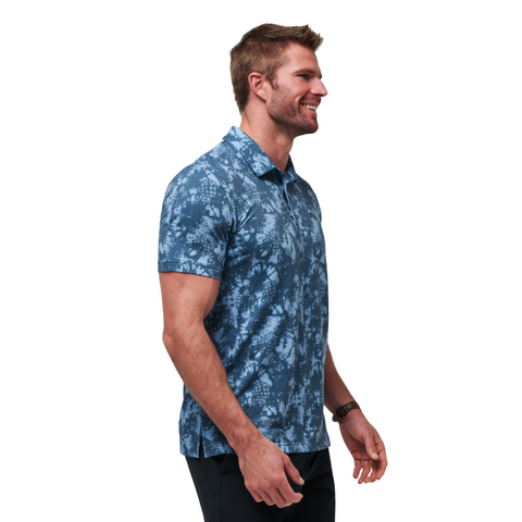 A smiling man with short brown hair and a beard wears a Travis Mathew Featherweight Please Mens Polo, a blue patterned golf shirt, with dark pants, standing against a white background.