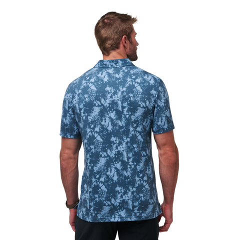 A man with short light brown hair stands with his back to the camera, wearing the Travis Mathew Featherweight Please Mens Polo in blue with a light blue floral pattern and black pants—ideal as a mens golf polo for moisture-wicking comfort.