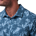 Close-up of a man wearing the Travis Mathew Featherweight Please Mens Polo in blue, featuring abstract shapes, collar and buttons, with an embroidered logo on the chest. His face is partially out of frame.