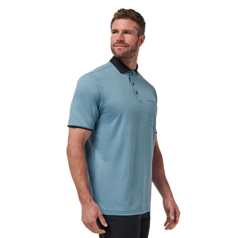 A man with short brown hair and a beard wears the Travis Mathew Featherweight Pocket Mens Polo in light blue with a black collar and sleeve trim, made from ultra-light performance fabric, standing and looking left against a white background.