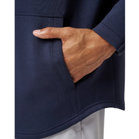 A close-up shows a hand partially inside the front pocket of the TravisMathew Fibercloud Men's Hoodie in Total Eclipse, with the sleeve visible and light-coloured pants.