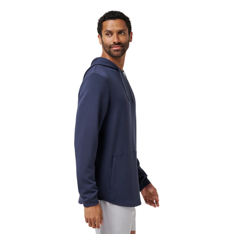 A man with short curly hair and a beard wears the TravisMathew Fibercloud Men's Hoodie in Total Eclipse, paired with white shorts. He stands sideways, looking over his shoulder and smiling against a white background.