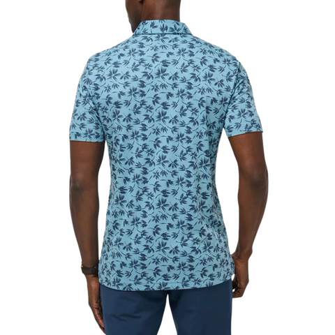 A man is seen from behind wearing a light blue Travis Mathew Jungle Polo Shirt featuring a dark blue palm leaf pattern, paired with navy blue pants.