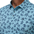 A close-up of a person wearing the Travis Mathew Jungle Polo Shirt, featuring a light blue color, dark blue leaf pattern, two buttons, and an embroidered chest logo.