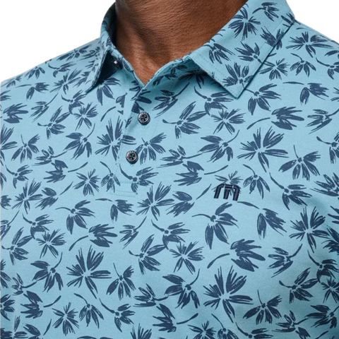 A close-up of a person wearing the Travis Mathew Jungle Polo Shirt, featuring a light blue color, dark blue leaf pattern, two buttons, and an embroidered chest logo.