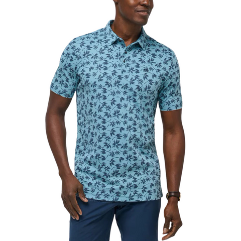 A man in a light blue Travis Mathew Jungle Polo Shirt with a dark blue palm tree pattern, dark blue pants, and a brown watch stands in a relaxed pose against a white background.