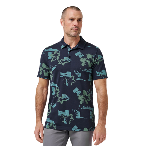 A man with short hair and a trimmed beard wears the Travis Mathew Men’s Favourite Hello Polo in Total Eclipse Blue, paired with light gray pants, standing against a white background.