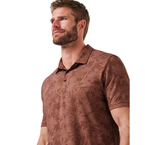A man with light brown hair and a beard wears the TravisMathew Mens Featherweight Ticket Polo Shirt in Maple, featuring a subtle leaf pattern. He looks up and left with a neutral expression against a white background.
