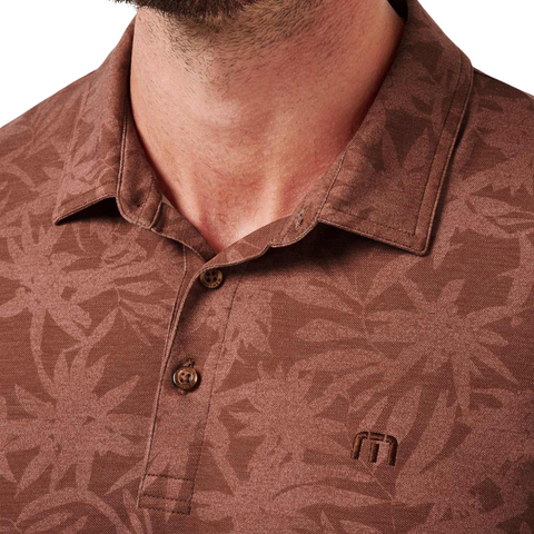 Close-up of a person wearing the Travis Mathew Men's Featherweight Ticket Polo Shirt in Maple, featuring a subtle floral pattern, embroidered logo, neckline detail, buttons.