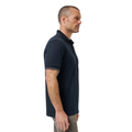 A man with short hair and a trimmed beard stands in profile, wearing a Travis Mathew Mens Featherweight Ticket Polo Shirt in Total Eclipse Navy and light-colored pants, against a plain white background.