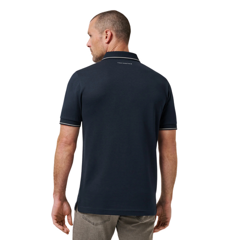 A man faces away wearing the Travis Mathew Mens Featherweight Ticket Polo Shirt in Total Eclipse Navy, featuring short sleeves, white trim, light gray pants, and small white text at the back of the neck.